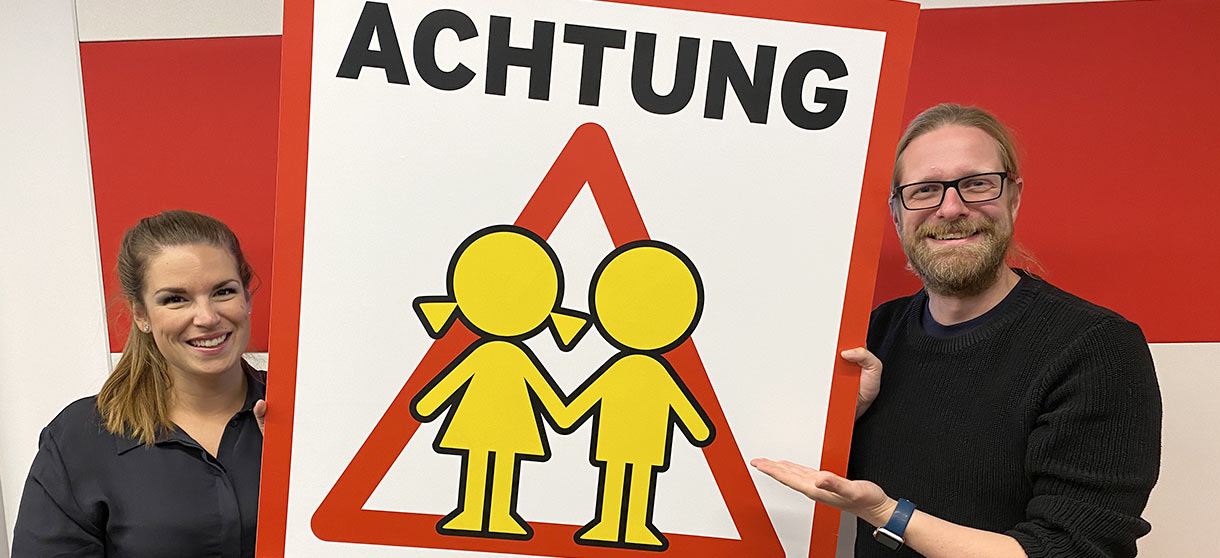 Die Guten-Morgen-Macher Carmen und Axel zeigen ein Achtung Kinder Plakat 
