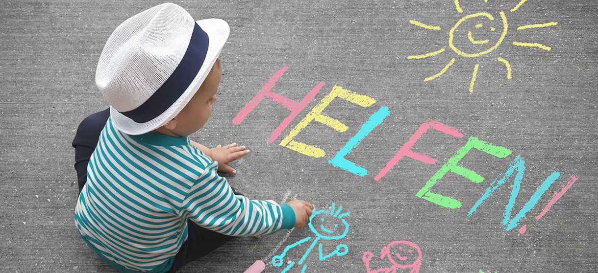 Kind schreibt mit bunter Kreide das Wort "Helfen" auf eine Straße